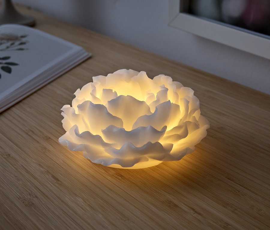 Bílá lampa ve tvaru květu svítí na dřevěném stole. Vedle ní leží otevřená kniha.