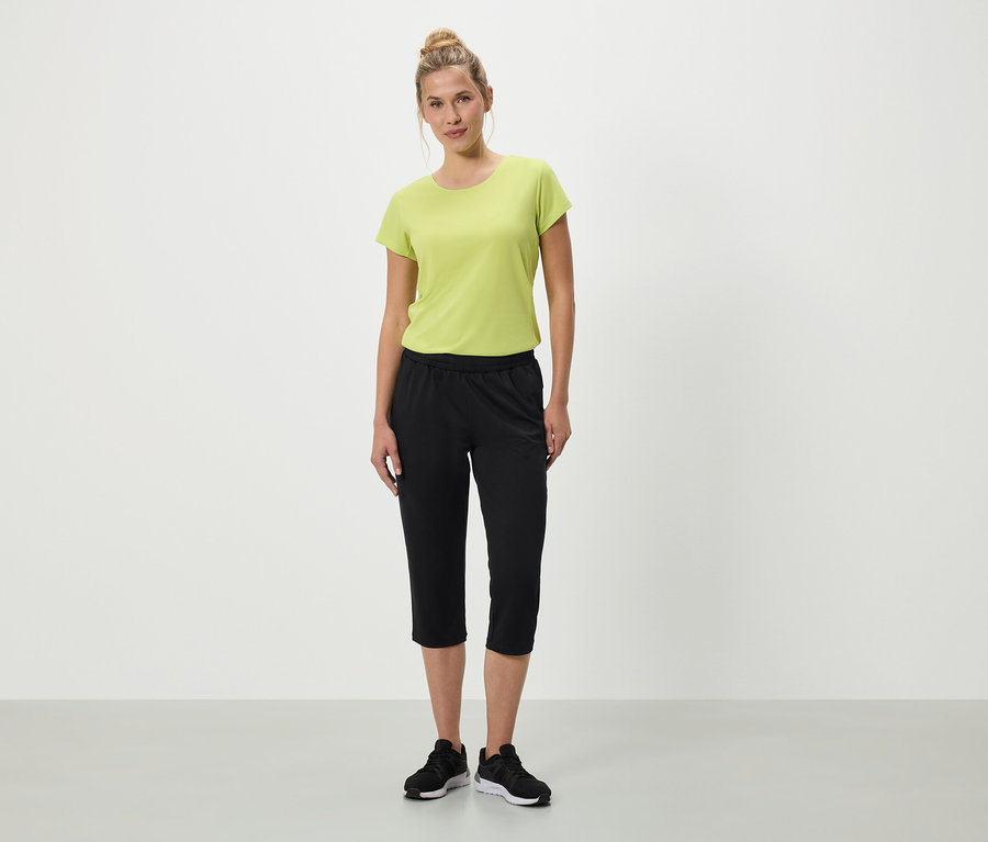 Žena stojí na bílém pozadí v zeleném tričku, černých tříčtvrtečních kalhotách a černých teniskách.