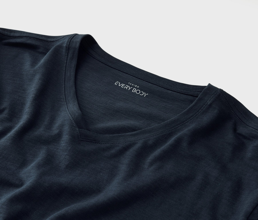Detailní záběr na tmavě modré tričko s véčkovým výstřihem a nápisem „Tchibo EVERY BODY“ pod límcem.