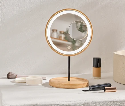 Na polici stojí kulaté, osvětlené kosmetické zrcadlo, make-up a štětec.