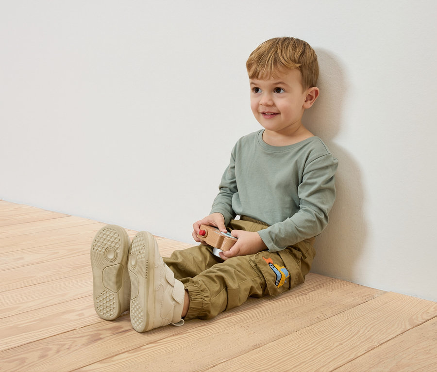 Usmívající se chlapec sedí na dřevěné podlaze, opřený o bílou zeď, a drží dřevěné autíčko.