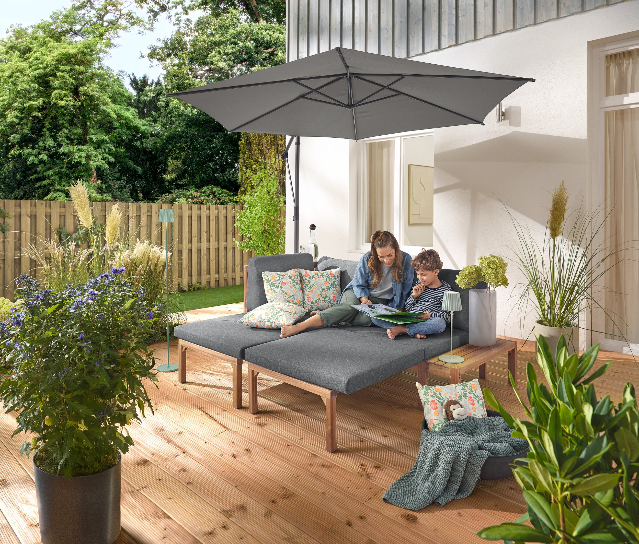 Žena a dítě sedí na šedé zahradní pohovce se samostatně výsuvnými podnožkami. Na pohovce leží dva venkovní polštáře s květinovým vzorem. Antracitový slunečník Leco se stojanem a krytem poskytuje stín. Na dřevěném stolku vedle pohovky stojí šalvějová venkovní dobíjecí stolní lampa LED a v pozadí je vidět šalvějová venkovní dobíjecí stojací lampa LED.