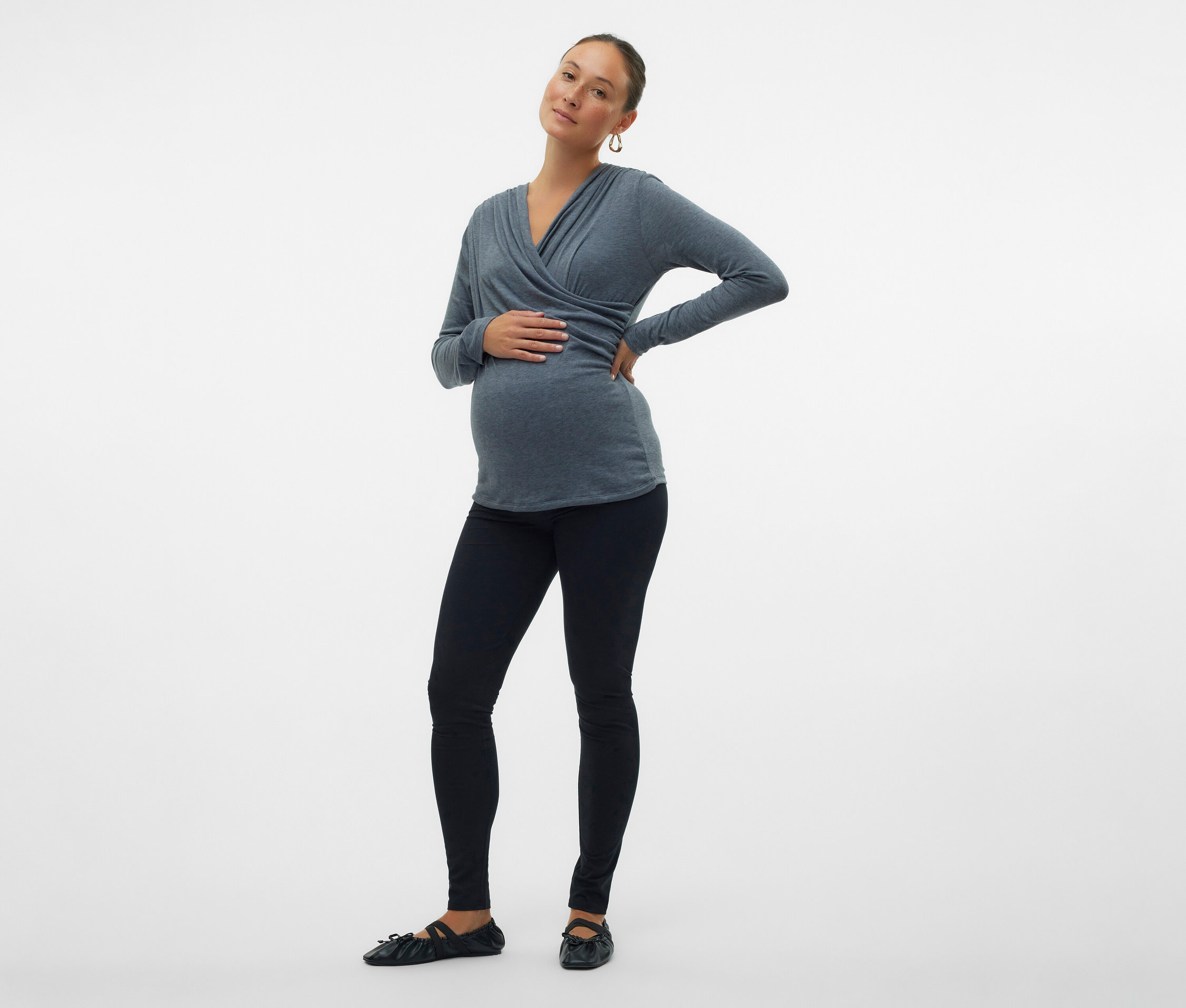 Těhotná žena v šedém zavinovacím topu, černých legínách a balerínkách pózuje na bílém pozadí.