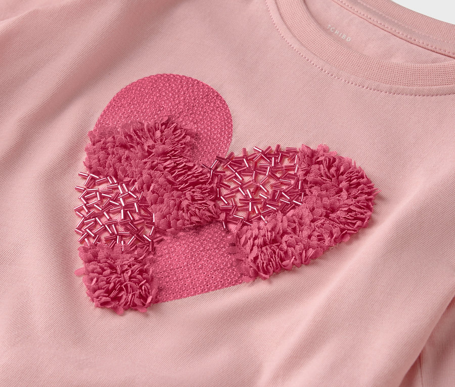 Detailní pohled na růžové tričko s nášivkou ve tvaru srdce zdobenou třásněmi a korálky.