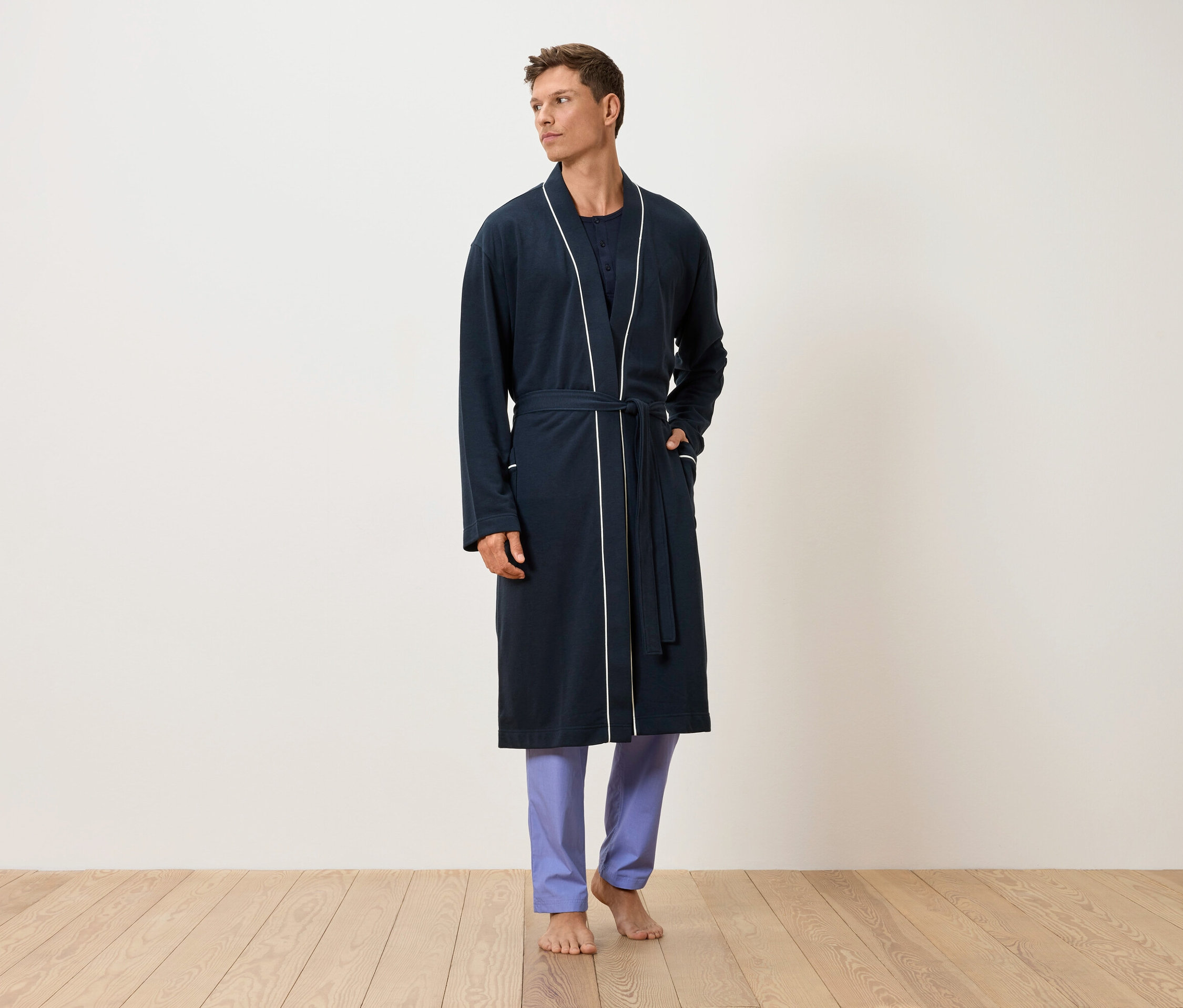 Muž stojí bosý na dřevěné podlaze, oblečený v tmavě modrém županu s bílým lemováním a modrých kalhotách.