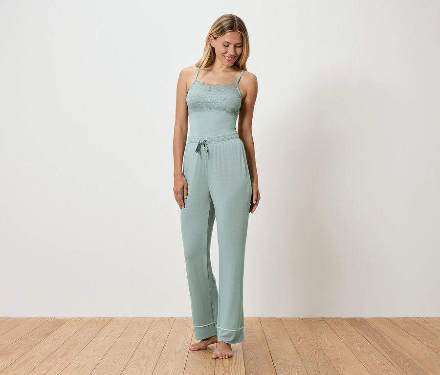 Žena má na sobě světle zelené pyžamo s krajkovými detaily, stojí na dřevěné podlaze před bílou stěnou.