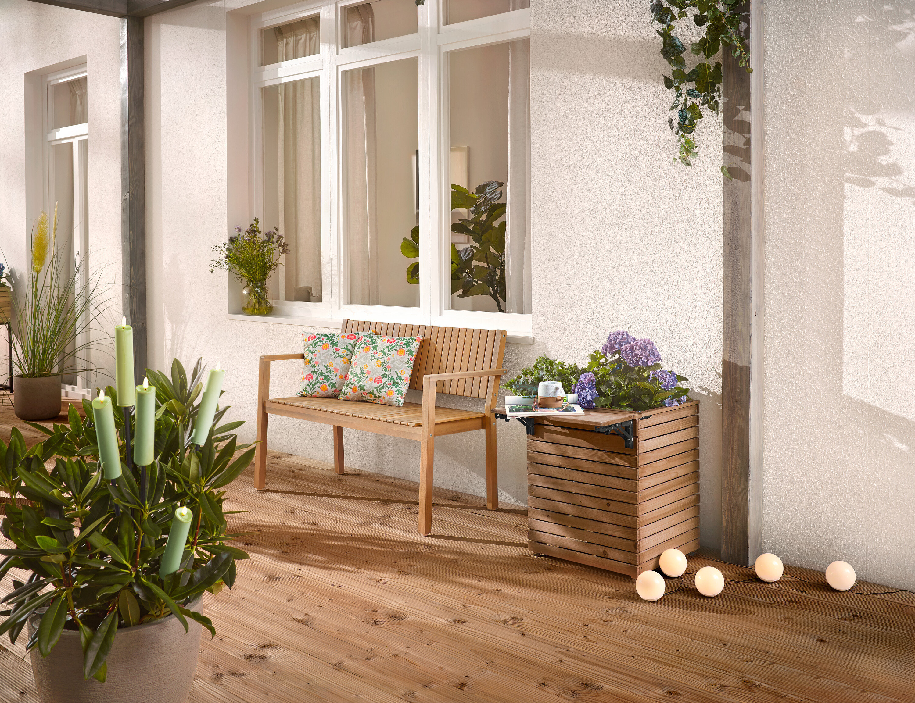 Na dřevěné terase stojí dřevěná lavice s polštáři, truhlík s květinami a hrnková rostlina se svíčkami.