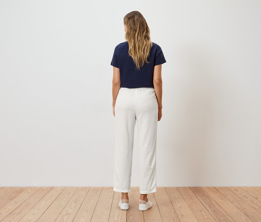 Žena s blond vlnitými vlasy stojí zády k divákovi, má na sobě tmavě modré tričko a bílé kalhoty.