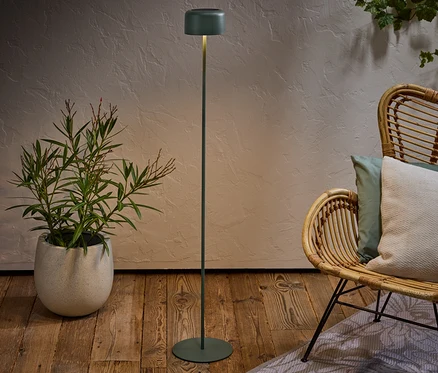Zelená stojací lampa stojí vedle ratanového křesla s polštáři a hrnkové rostliny na dřevěné podlaze.