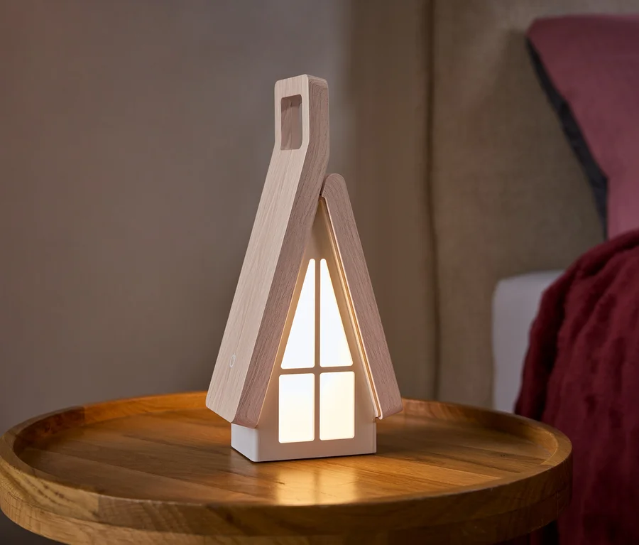 Lampa ve tvaru domečku se světlými dřevěnými boky a svítícími bílými okny stojí na kulatém dřevěném odkládacím stolku. V pozadí je postel s růžovým polštářem a červenou dekou.