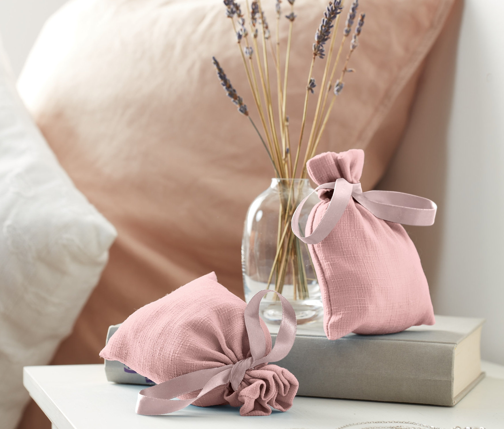 Dva vonné sáčky leží na knize vedle vázy s levandulí.