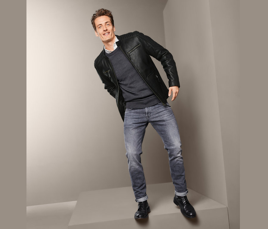 Muž pózuje v kožené bundě motorkářského stylu, svetru a džínách.