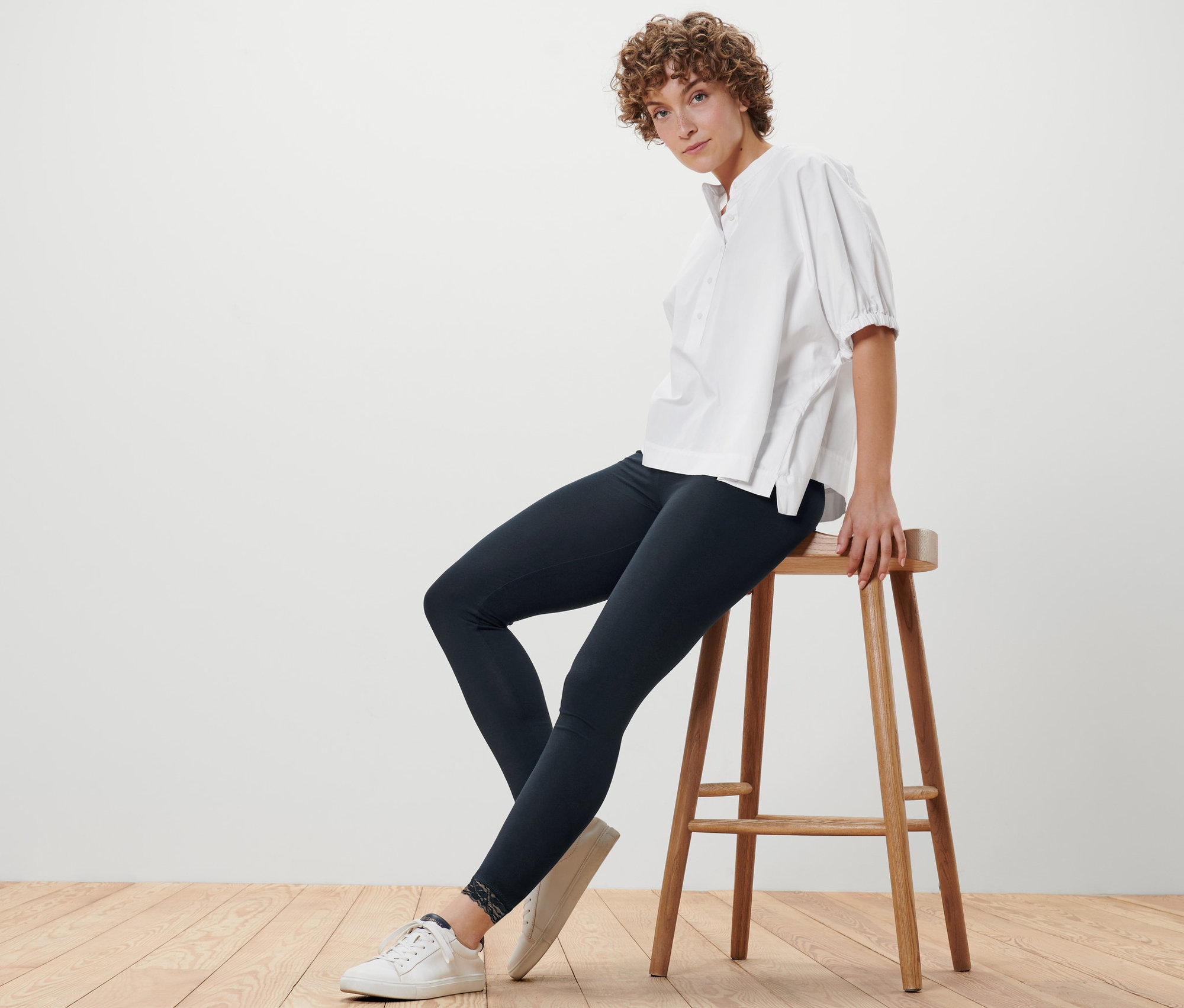Žena sedí na stoličce v černých legínách s krajkovým lemem a bílé blůze.