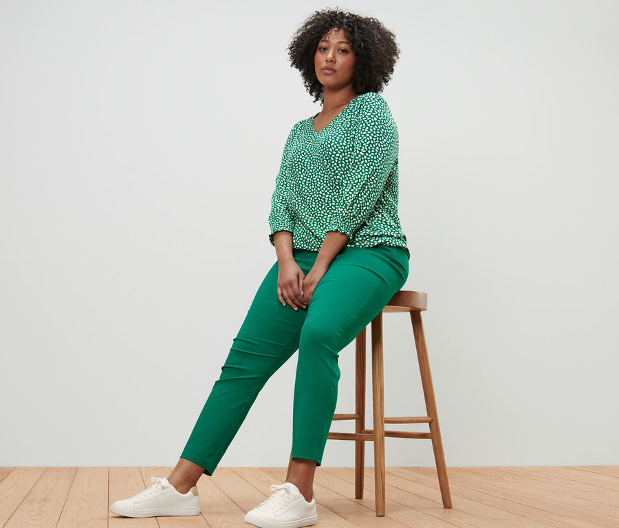 Žena sedí na stoličce a má na sobě zelené halenkové triko s potiskem a zelené strečové kalhoty.