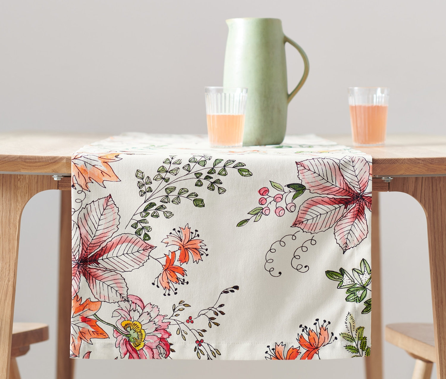Na dřevěném stole je běhoun s květinovým vzorem. Na stole stojí džbán a dvě sklenice s oranžovou tekutinou.
