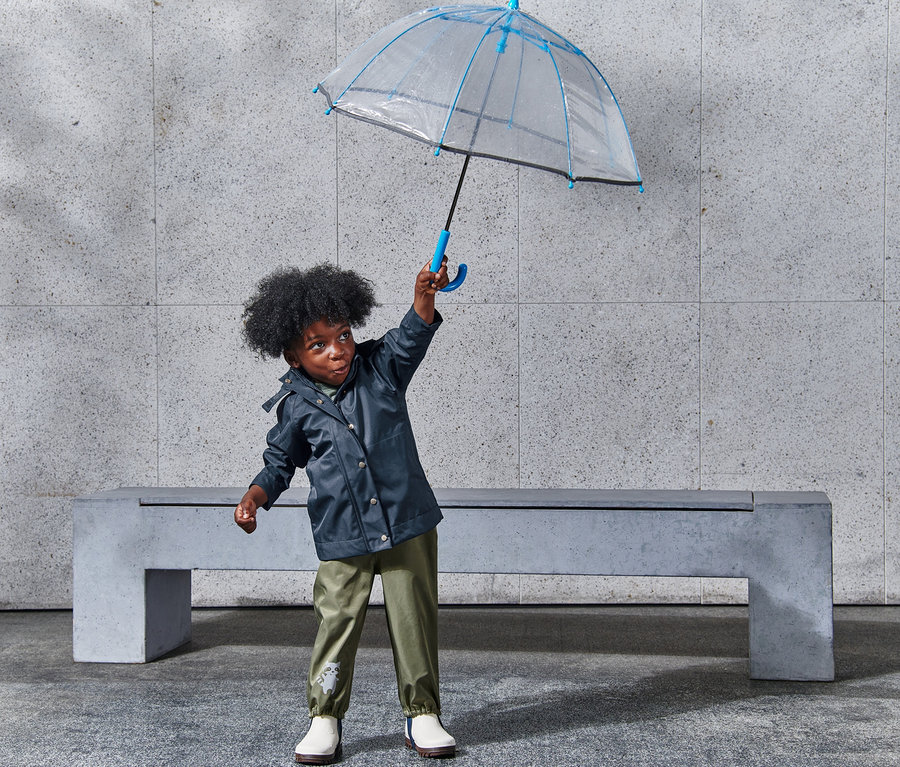 Chlapec drží nad hlavou průhledný deštník s modrým lemováním a má na sobě bundu do deště.