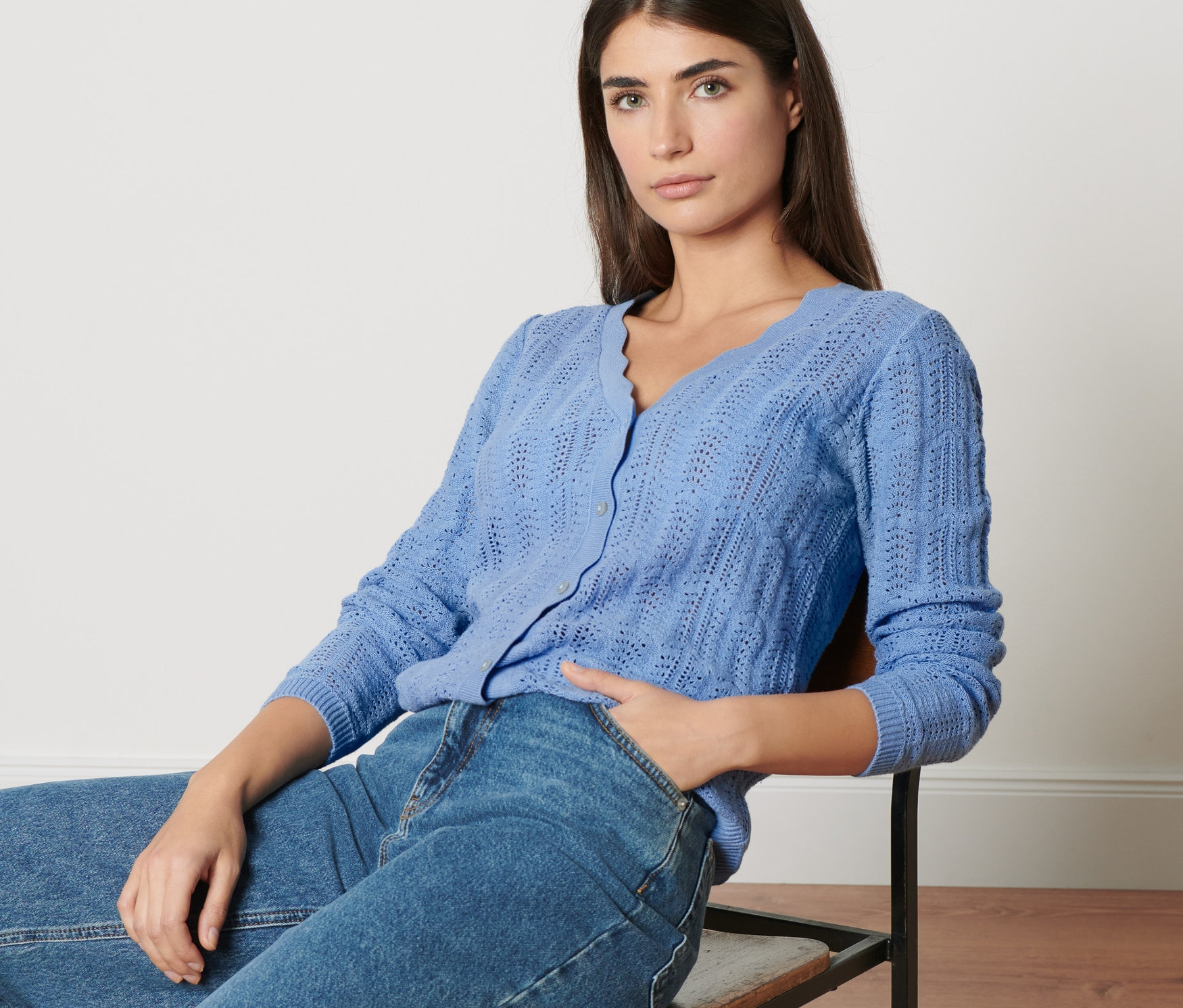 Žena sedí na židli a má na sobě pletený kabátek s ažurovým vzorem a džíny.