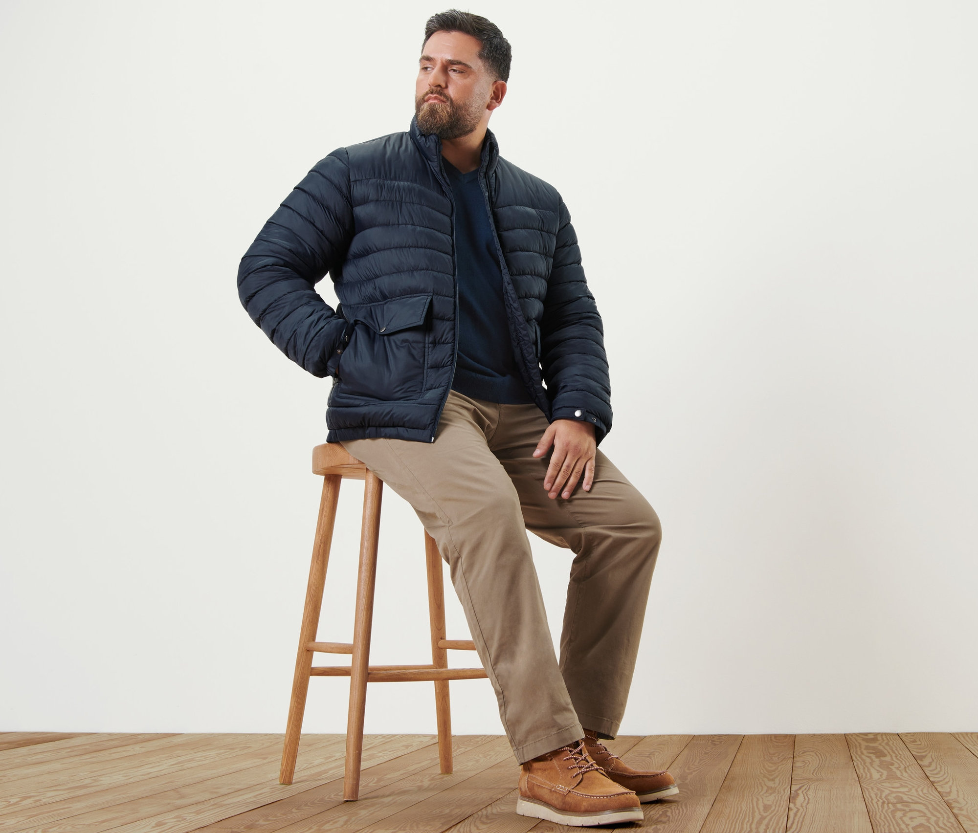 Muž sedí na stoličce a má na sobě pískové chino kalhoty, tmavomodrý svetr s výstřihem do V a námořnicky modrou prošívanou bundu.