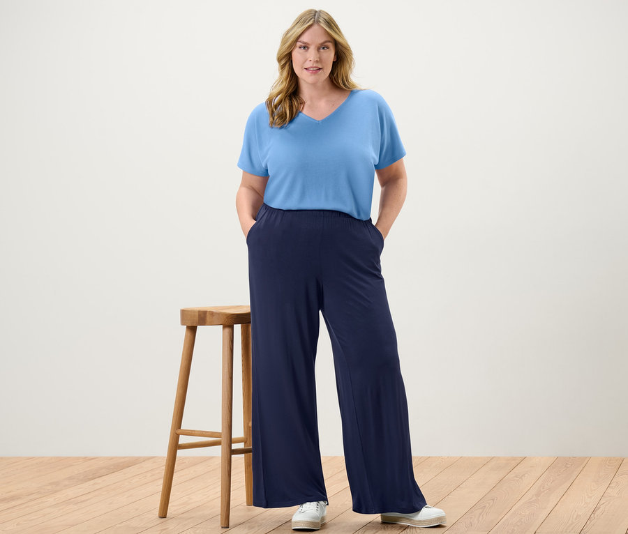 Žena stojí a má ruce v kapsách tmavě modrých kalhot se širokými nohavicemi a světle modrého strukturovaného trička.