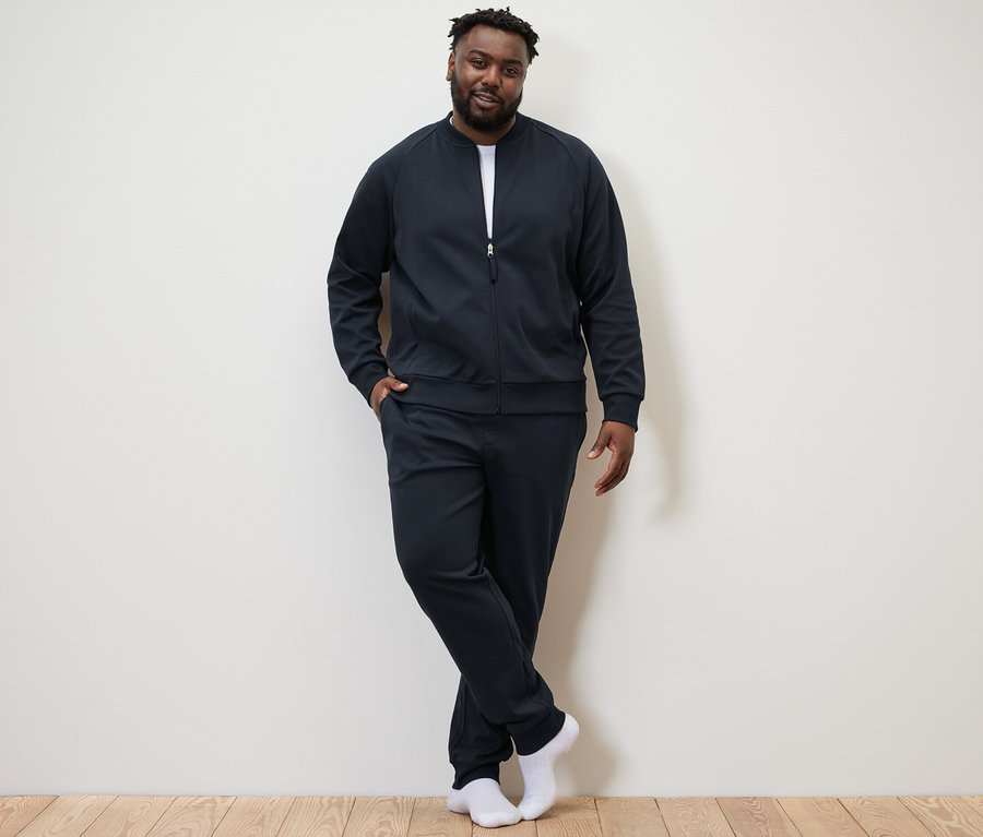 Muž pózuje na pozadí zdi a má na sobě tmavě modrou domácí soupravu.