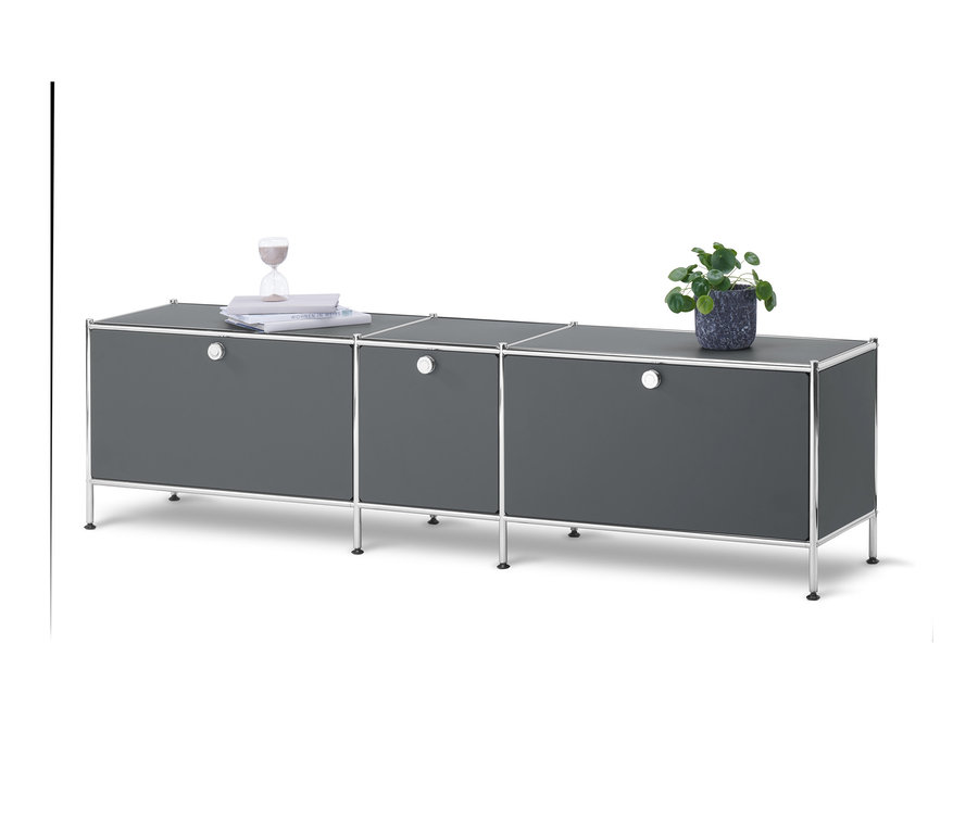 Šedá nízká skříňka CN3 se 3 výklopnými dvířky stojí na bílém pozadí. Na skříňce je rostlina v květináči, knihy a přesýpací hodiny.