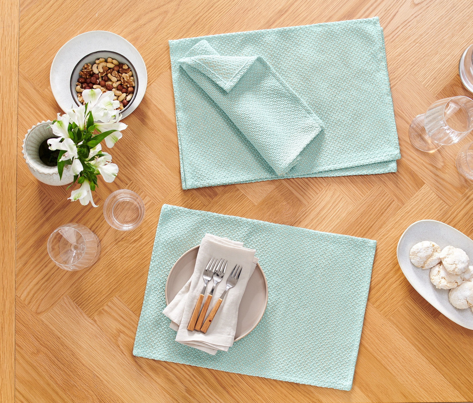 Dva světle modré prostírání a odpovídající ubrousek leží na dřevěném stole vedle talíře s ořechy, květin a sklenic.