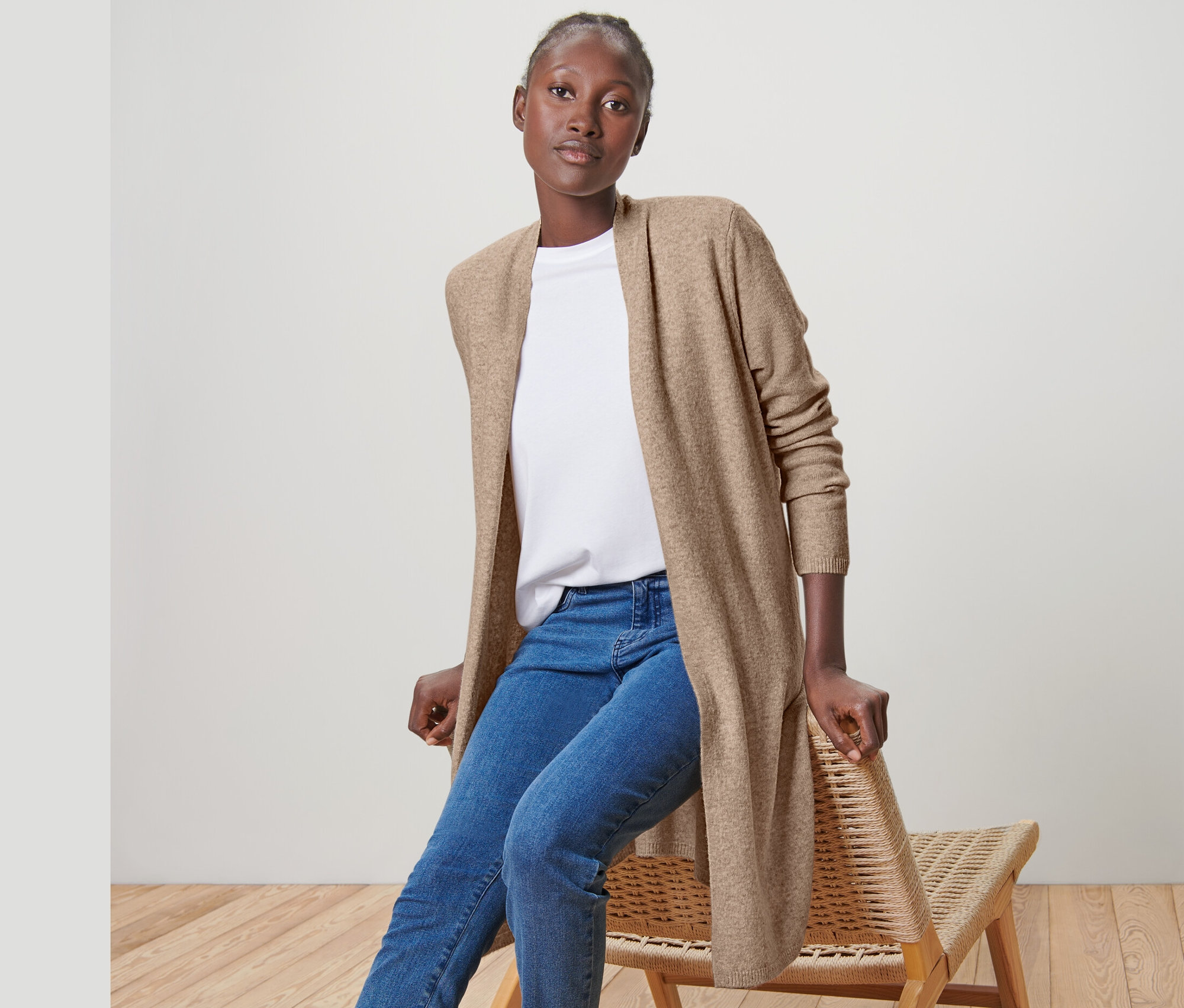 Žena sedí na židli a má na sobě otevřený dlouhý kardigan, džíny a tenisky.