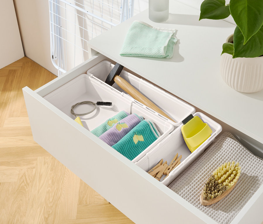 Otevřená zásuvka bílého úložného prostoru s různými předměty, včetně hadříků, kladiva a kartáče.