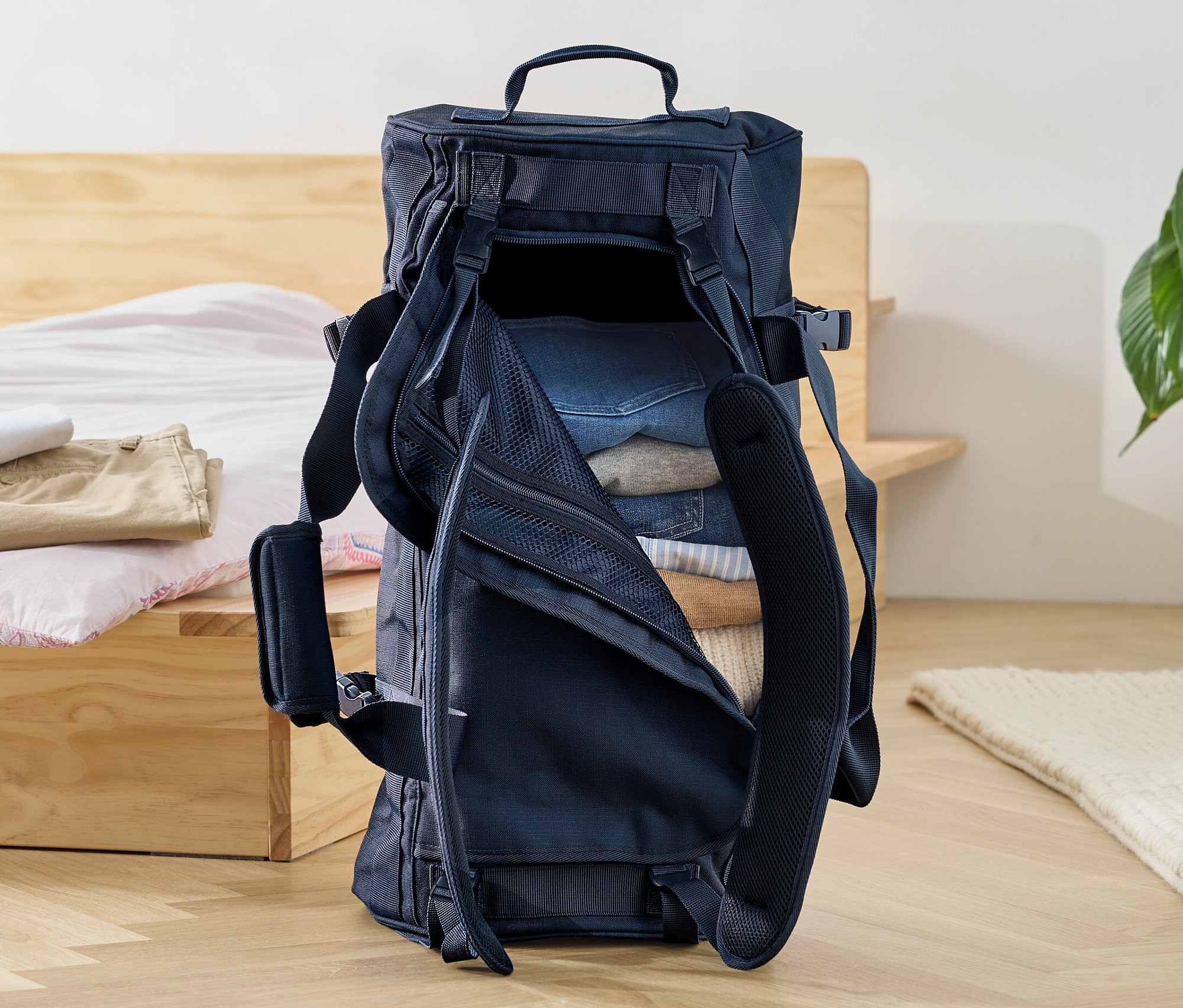 Otevřená cestovní taška a batoh naplněná oblečením stojí na podlaze u postele.