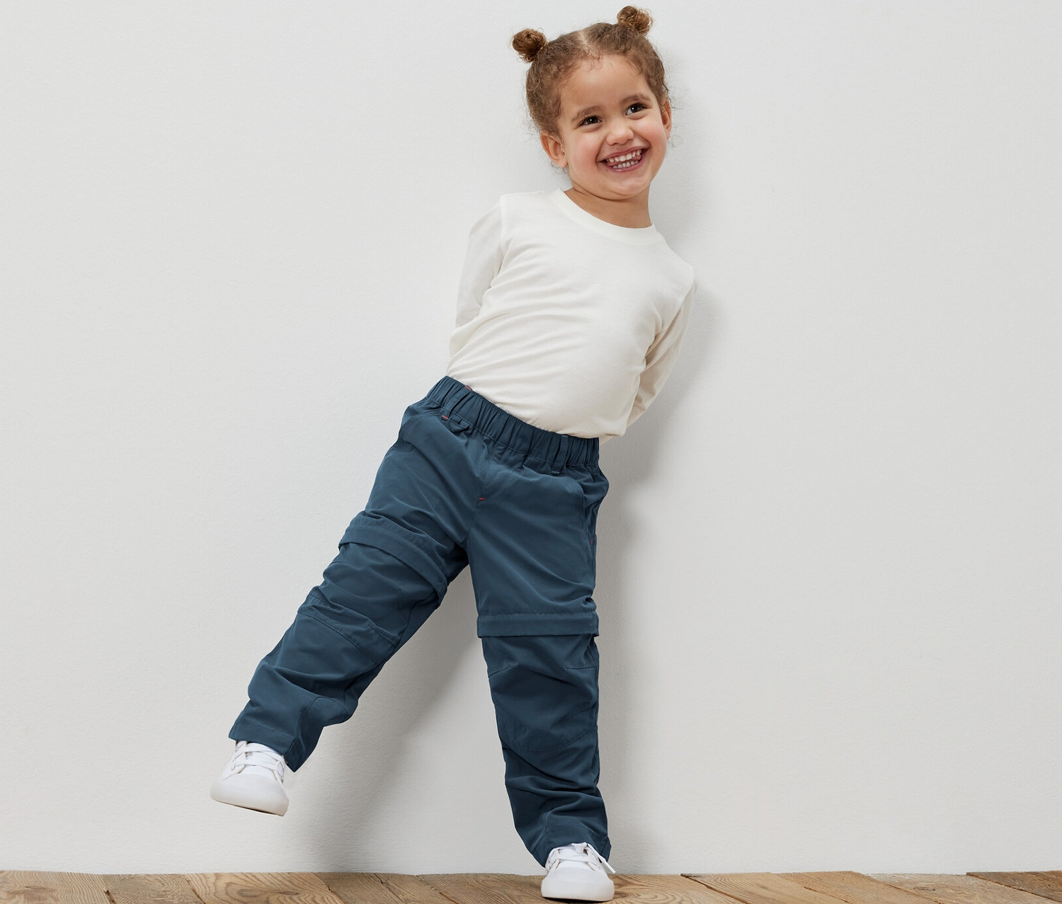 Usmívající se holčička opřená o bílou zeď má na sobě funkční kalhoty pro malé děti a bílou košili.