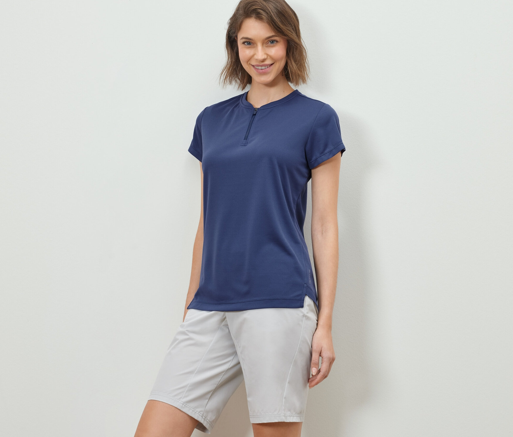 Žena pózuje ve sportovním triku s polovičním zipem a šortkách.