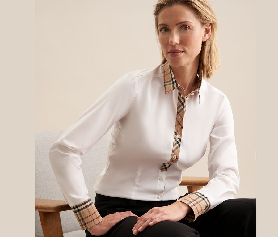 Žena sedí na židli a má na sobě bílou popelínovou halenku Seidensticker s kostkovanými detaily.