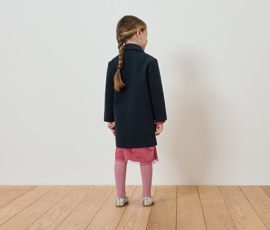 Dívka zády k fotoaparátu má na sobě tmavě modrý kabát, růžové šaty a punčochy a stříbrné boty.