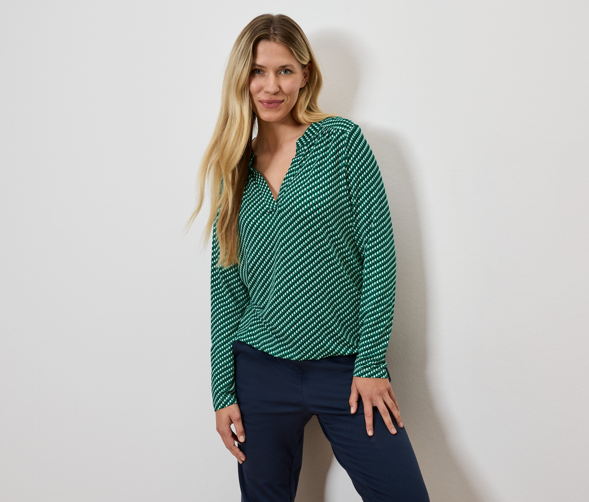 Žena s blond vlasy pózuje v zelené halence s proužky a tmavě modrých kalhotách.