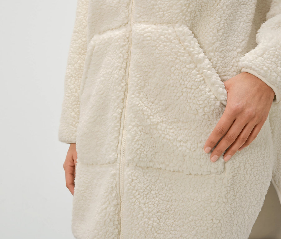 Detail dámského krémového županu z umělé kožešiny se zipem a kapsami, ruka v kapse.