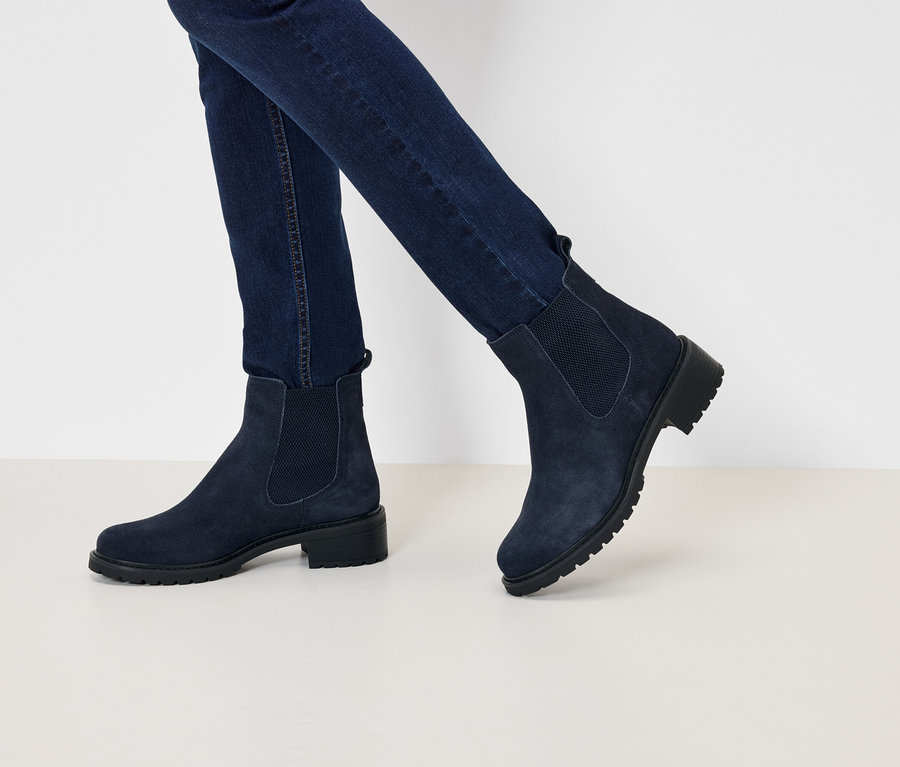 Nohy v tmavě modrých džínách a semišových botách Chelsea na nízkém podpatku, kráčející po bílém povrchu.