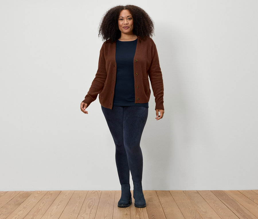 Žena s hnědými kudrnatými vlasy stojí před bílou zdí a má na sobě hnědý kardigan, tmavě modré tričko, tmavě modré legíny a tmavě modré boty.