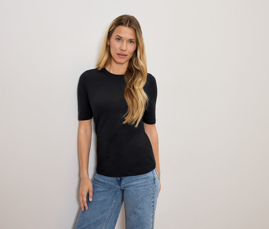 Žena s blond vlasy pózuje v černém tričku a modrých džínách.