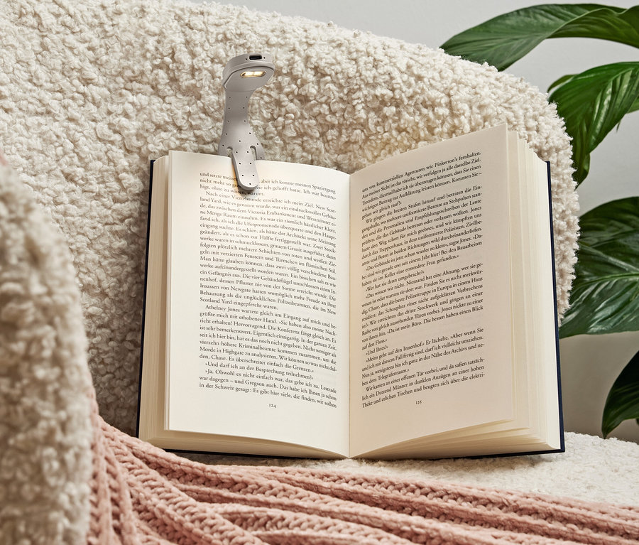 Šedá lampička na čtení je připevněna k otevřené knize ležící na měkké krémové dece s růžovými akcenty.