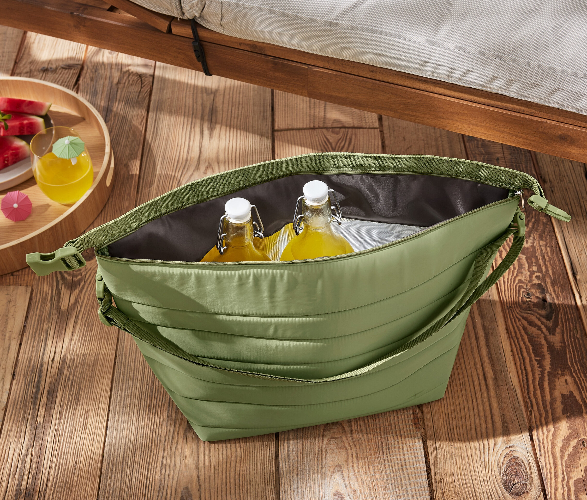 Zelená chladicí taška s lahvemi a melounem na dřevěném stole.