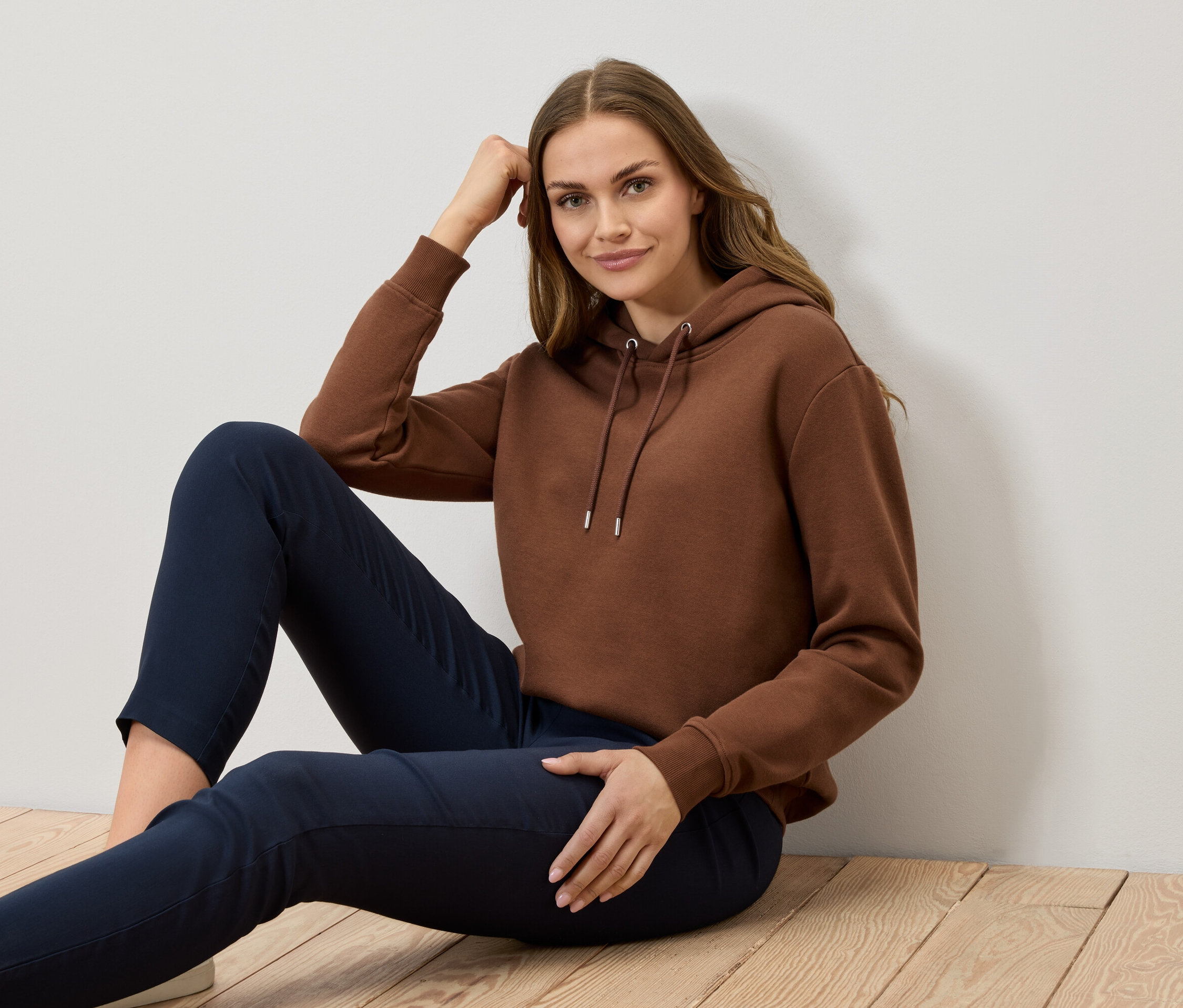 Žena sedí na dřevěné podlaze a má na sobě hnědou mikinu a tmavě modré kalhoty.