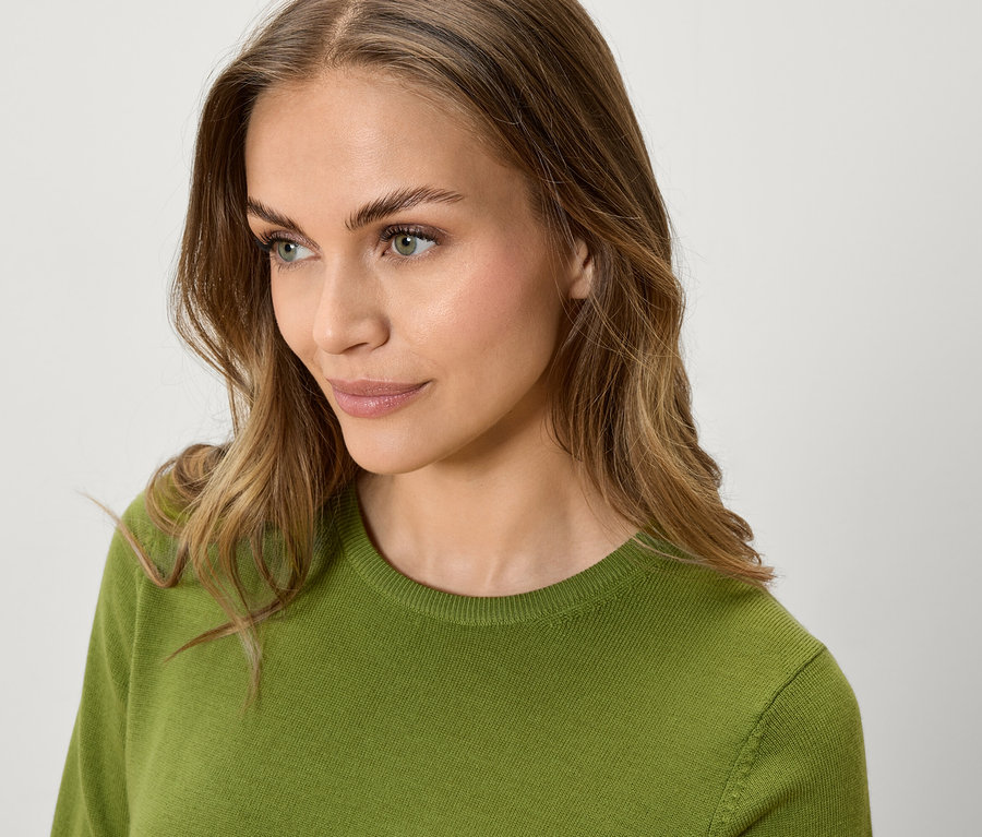 Portrét ženy s hnědými vlnitými vlasy a zeleným svetrem, dívá se do strany.