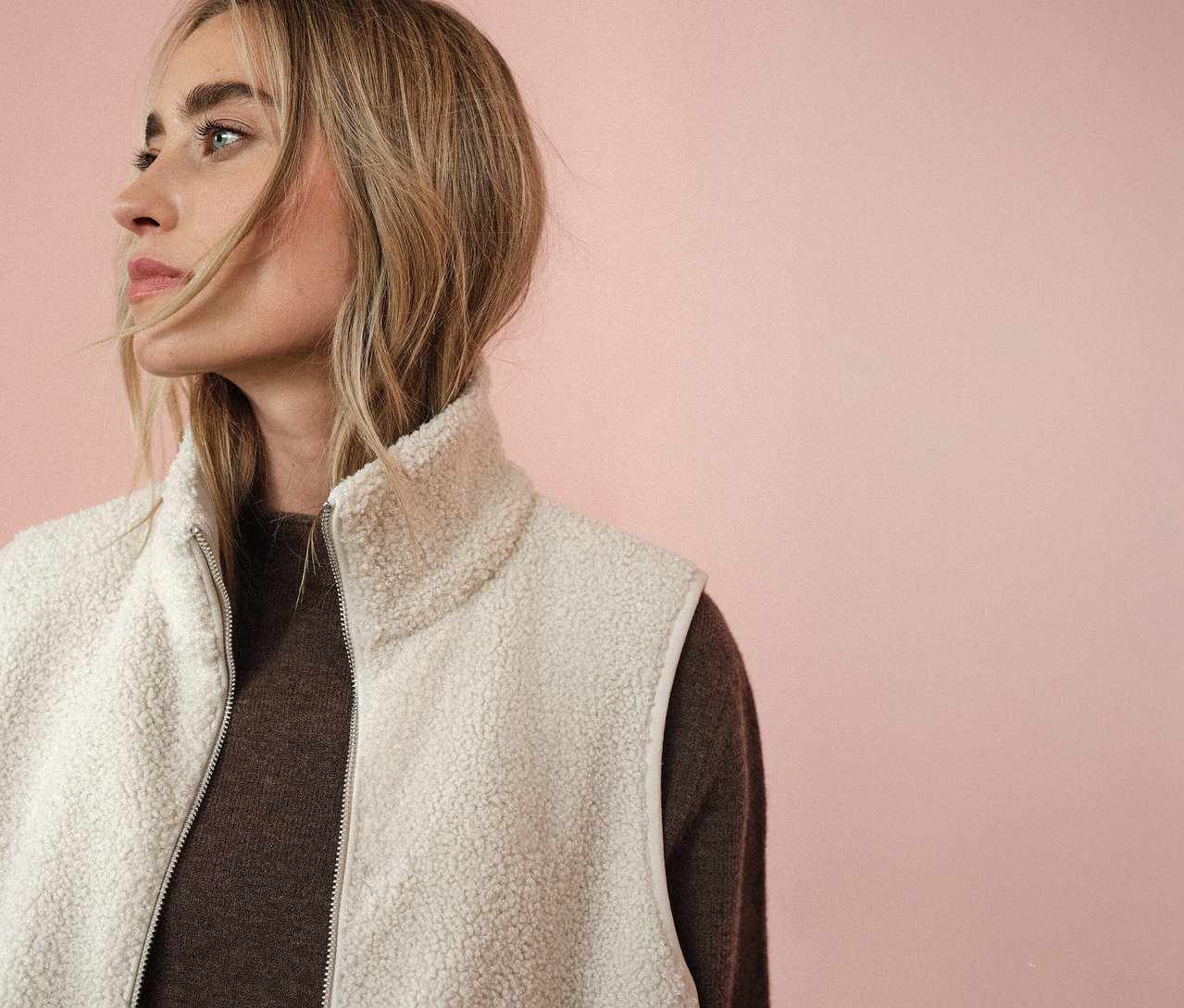 Žena nosí plyšovou vestu Street One přes hnědý svetr, dívá se doleva na růžovém pozadí.