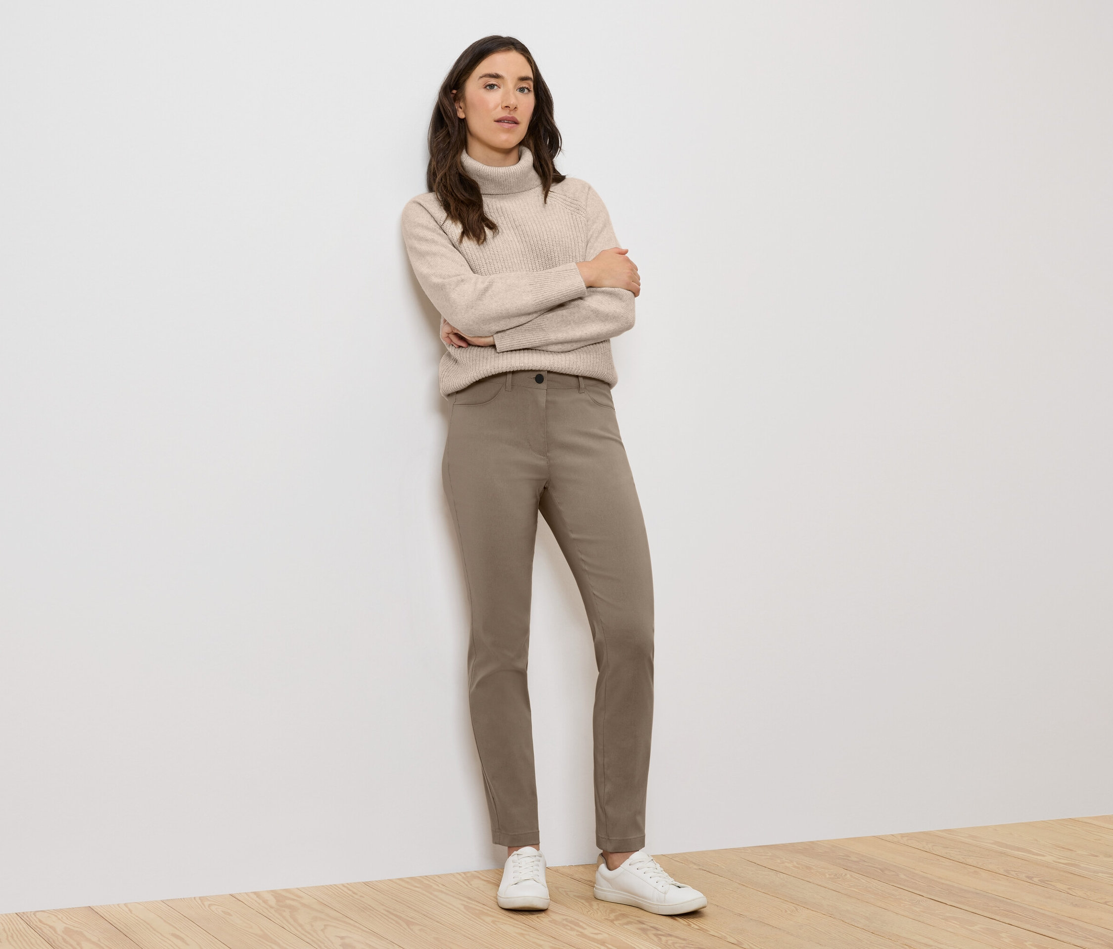 Žena stojí s rukama zkříženýma na prsou, má na sobě béžový rolák, hnědé kalhoty a bílé tenisky.