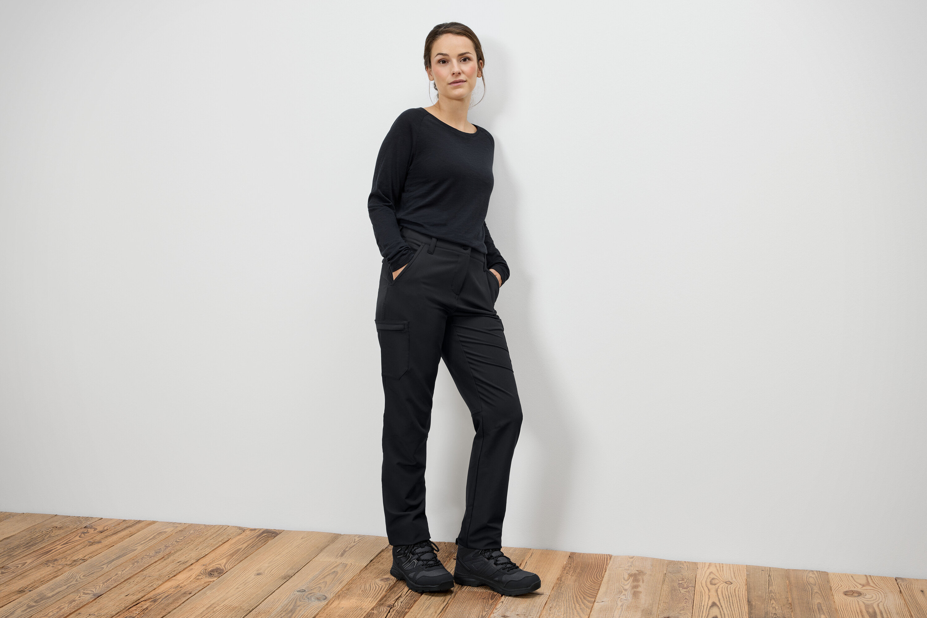 Žena stojí a má ruce v kapsách černých kalhot a černé boty.