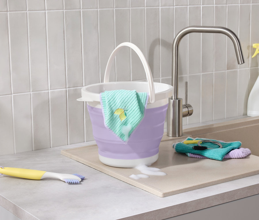 Fialový skládací kbelík s hadříkem s výšivkou citronu stojí na lince vedle dřezu, kde je žlutý kartáč a hadříky.
