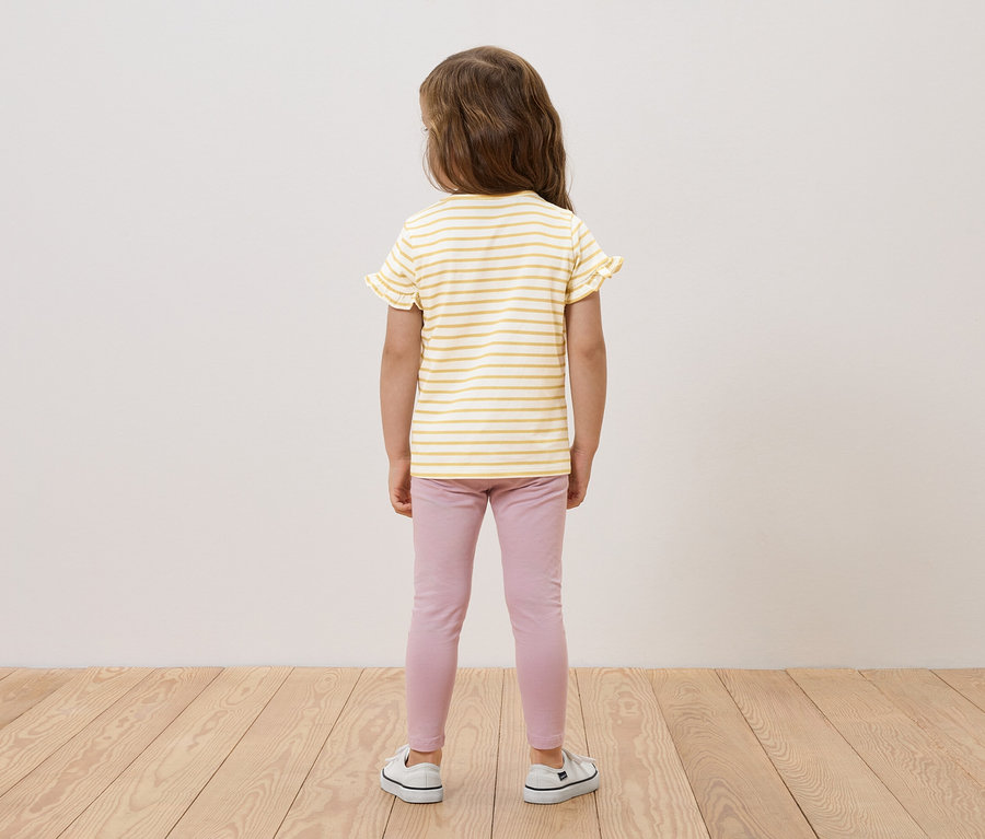Dívka stojí zády, má na sobě žluto-bílý pruhovaný top s volánky, růžové legíny a bílé tenisky.