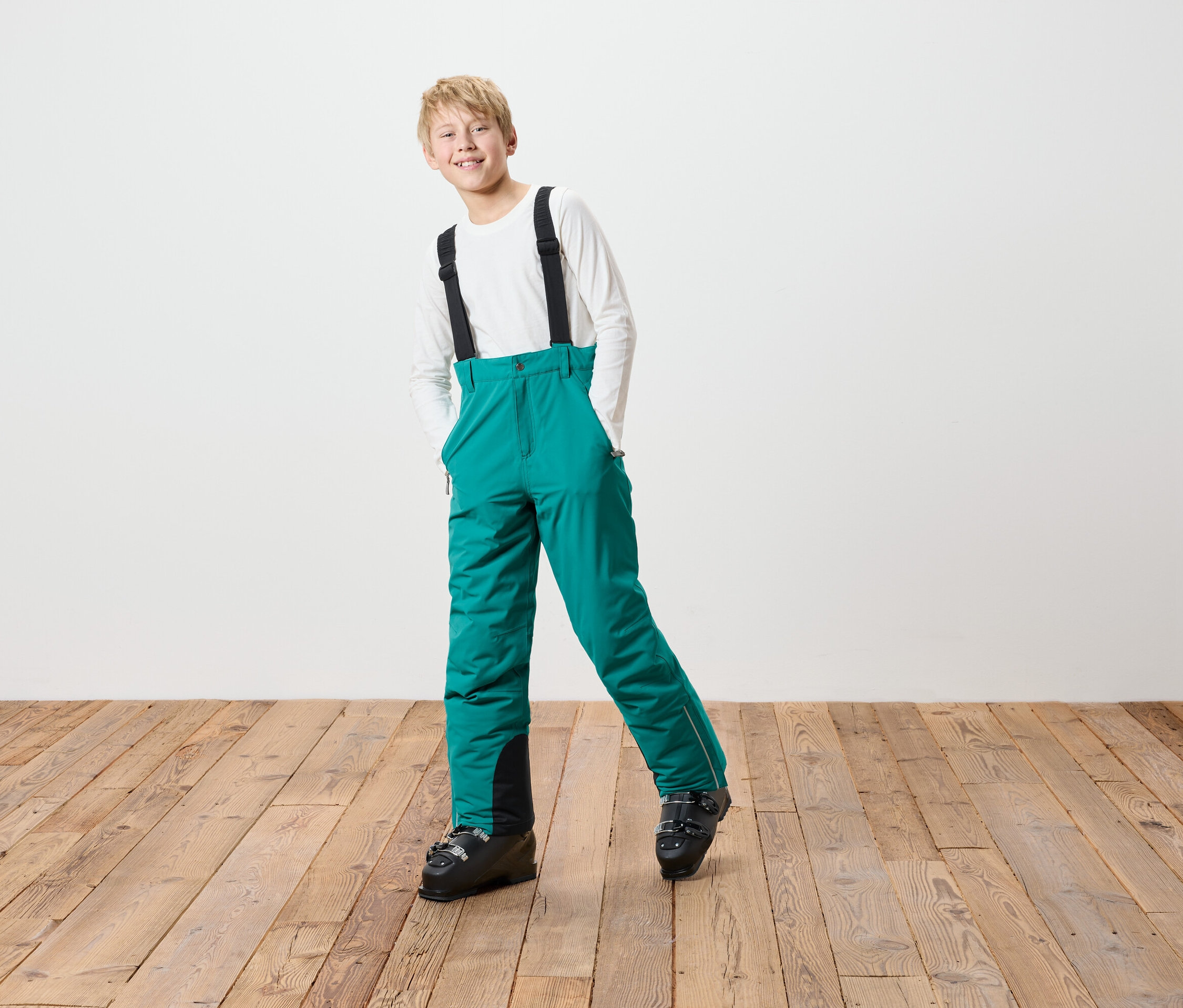 Chlapec v zelených lyžařských kalhotách s černými šlemi a lyžařských botách stojí na dřevěné podlaze před bílou zdí.