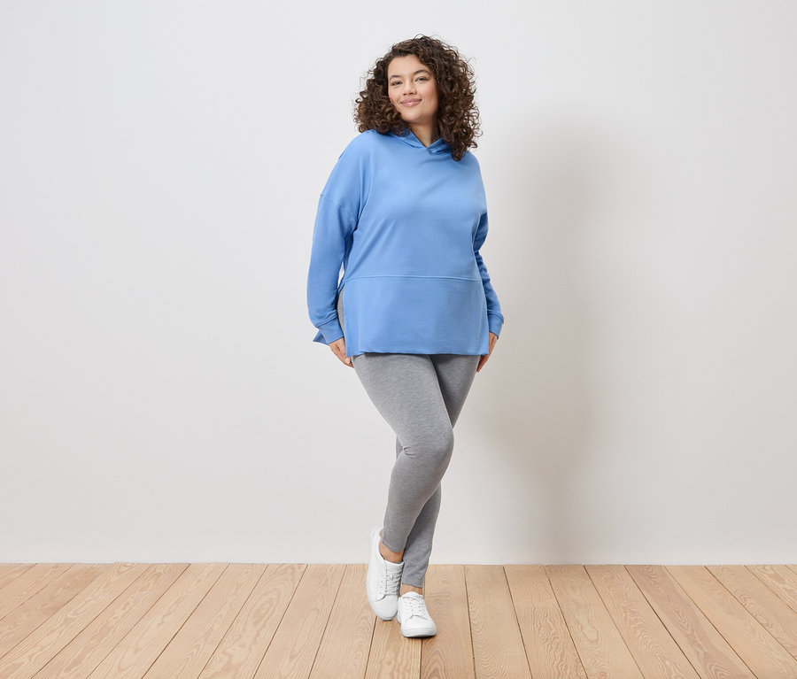 Žena s kudrnatými vlasy stojí před bílou zdí a má na sobě modrou mikinu s kapucí, šedé legíny a bílé tenisky.