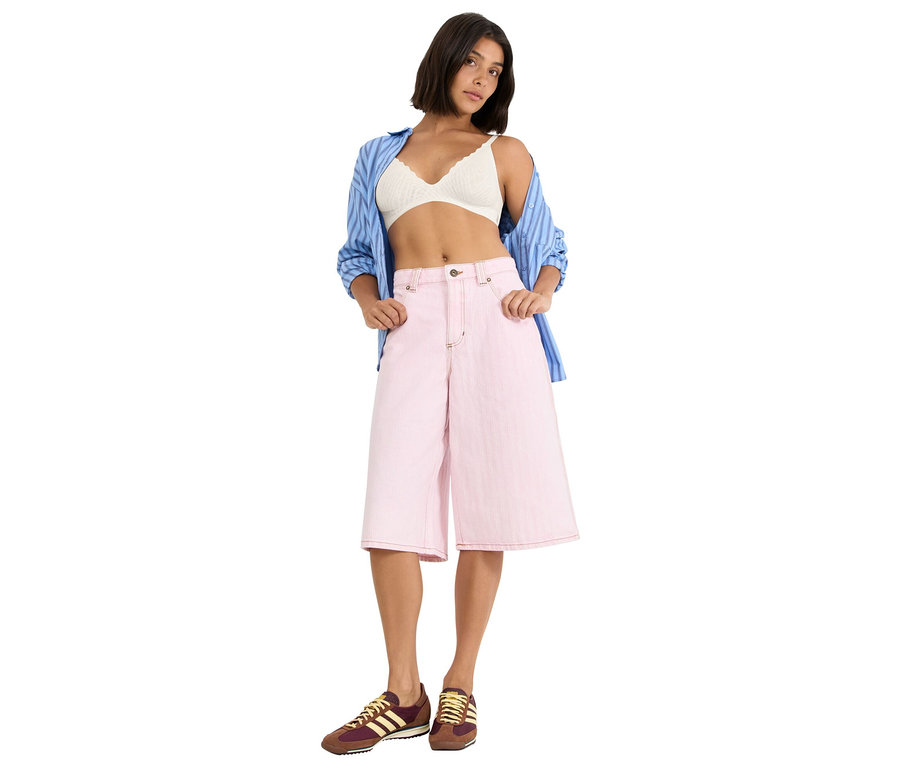 Žena má na sobě bílou krajkovou podprsenku, modrou košilovou halenku a růžové culottes kalhoty a hnědé a žluté tenisky.